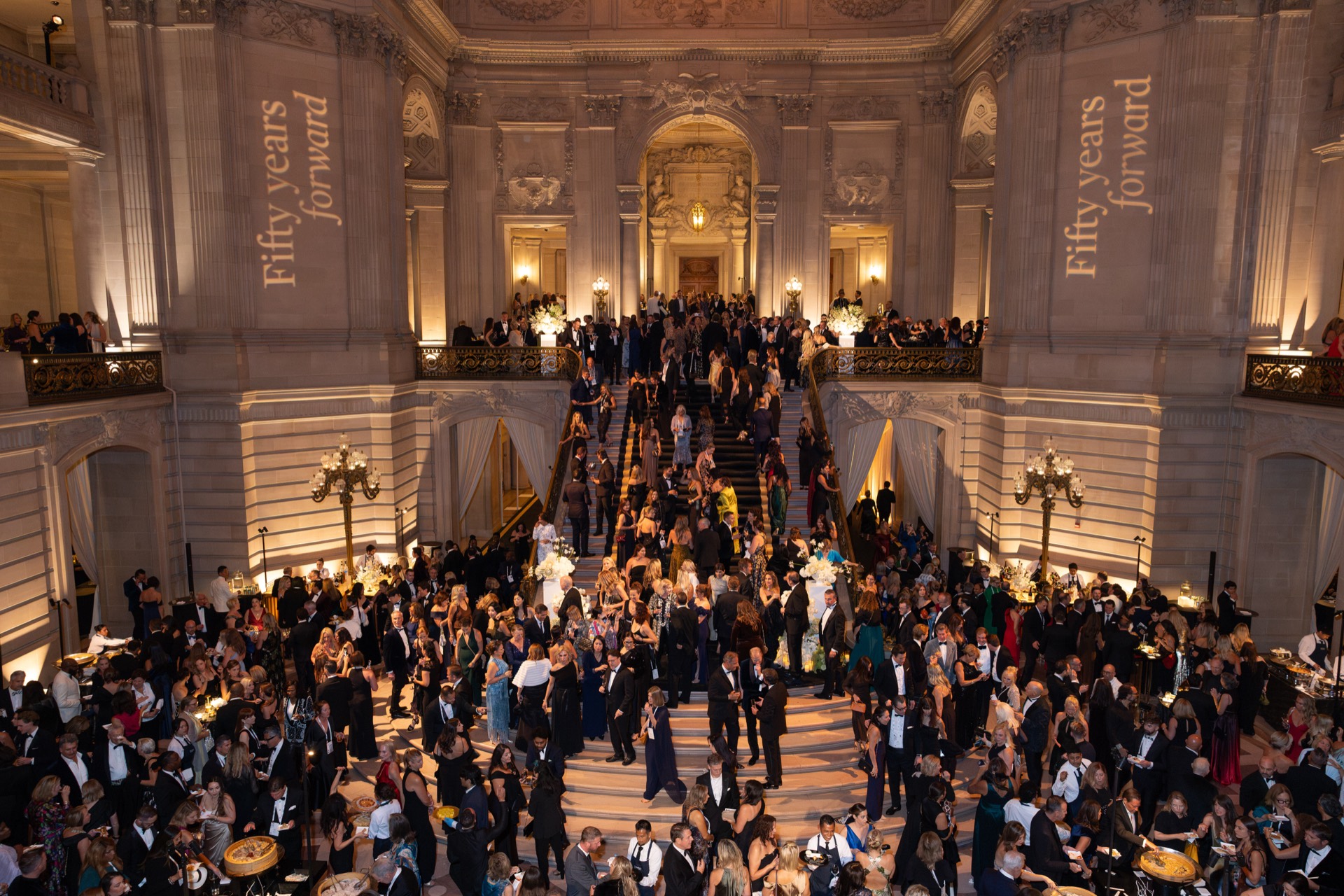 Guests in formal attire gather on the grand staircase at San Francisco City Hall beneath a “Fifty years forward” banner